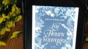Liseuse avec la couverture du livre d'Holly Ringland, Les fleurs Sauvages. Genêts en arrière plan