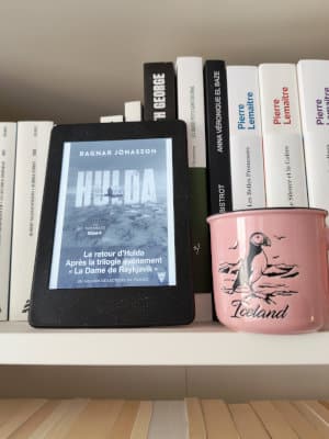 Liseuse posée sur la bibliothèque à côté d'un mug avec un macareux et la mention Iceland. La liseuse présente la couverture du livre de Ragnar Jonasson, Hulda