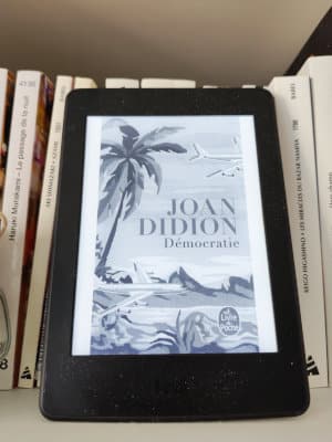 Liseuse posée sur une étagère de bibliothèque. Sur la liseuse, la couverture du livre de Joan Didion, Démocratie